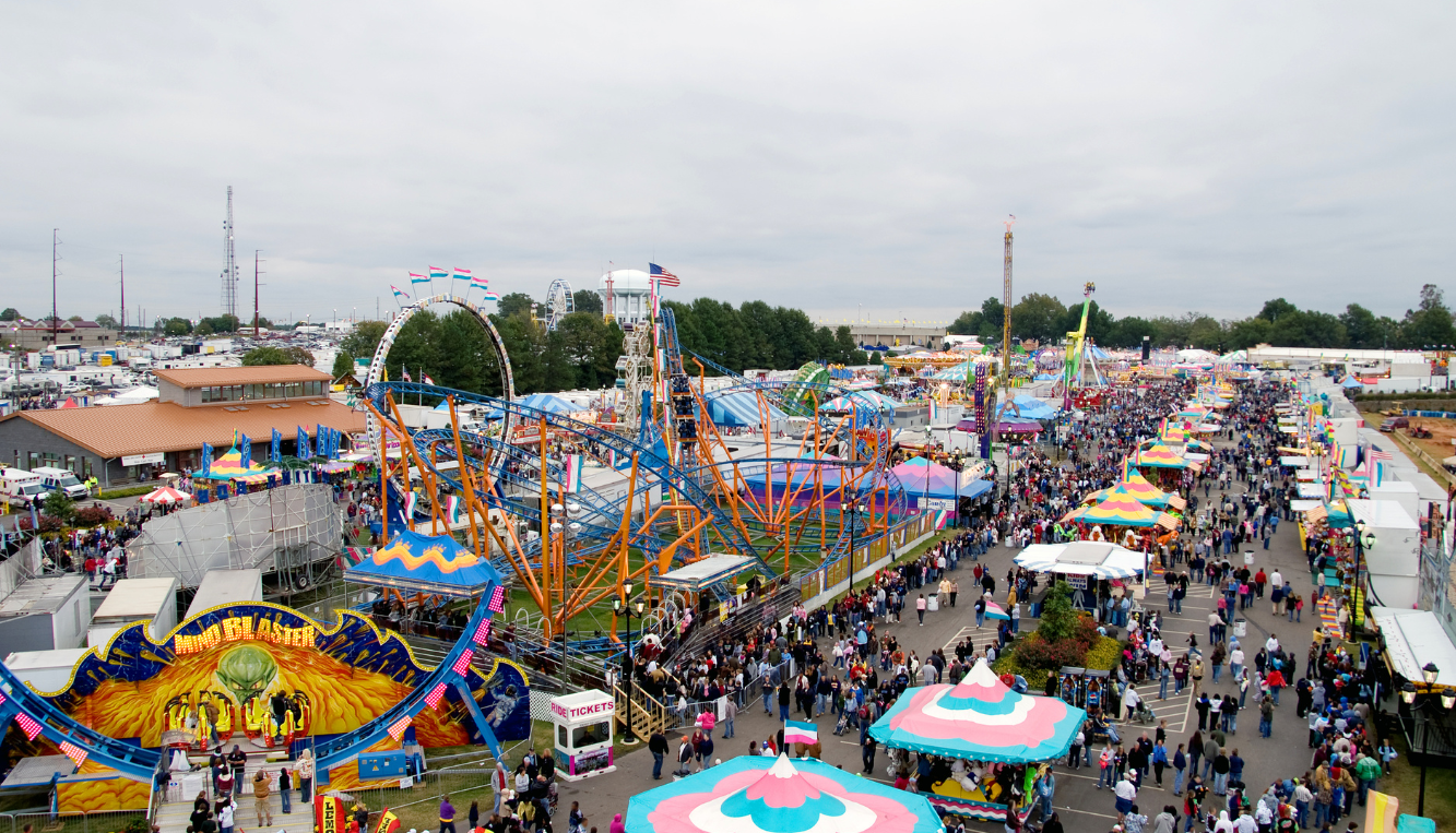 Fairground Aerial View Aerial view of a local fairground with various rides, including a large roller coaster, tents with colorful tops, and a crowd of people walking through the fair.