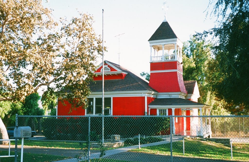 Santa Clara Schoolhouse, a red building with a prominent tower, set behind a fence, 1985.