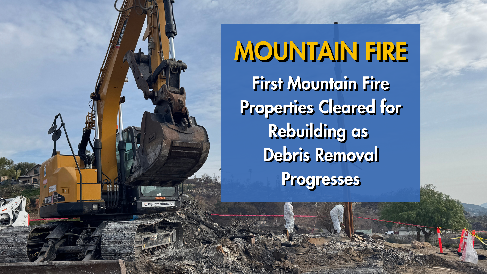 Excavator clearing debris with workers in protective gear nearby.