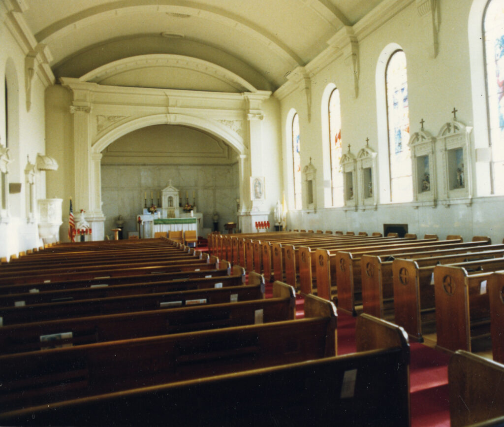 Interior of St. Mary Magdalen Church with wooden pews, stained glass windows, and barrel vault ceiling.